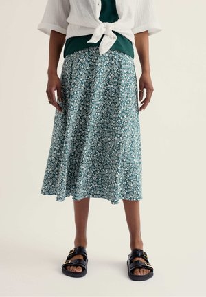 Személy türkiz virágos midi szoknyában, fehér megkötős ingben, sötétzöld felső fölött, fekete csatos szandálokkal, egy egyszínű háttér előtt áll.