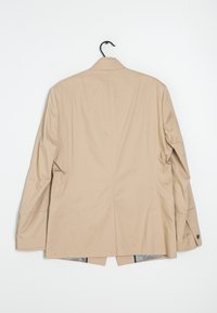 Beżowa marynarka wykonana z gładkiego materiału, z jednym rozcięciem z tyłu i mankietami zapinanymi na guziki. Wisi na czarnym haczyku na białym tle.