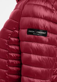 Czerwona pikowana kurtka puchowa z lśniącą, gładką fakturą. Zawiera czarną metkę z napisem "FRIEDA FREDDIES NEW YORK" na rękawie.