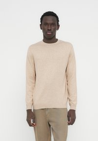 Pull beige à manches longues avec col rond, texture tricotée et poignets et ourlet côtelés, porté avec un pantalon beige clair.