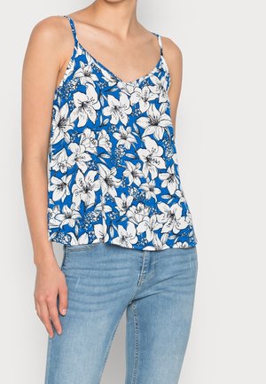 Frau trägt ein blaues ärmelloses Oberteil mit weißem Blumenmuster und hellblaue Jeans, steht vor einem einfarbigen hellen Hintergrund.