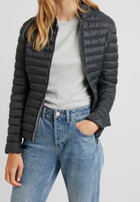 Sort quilted jakke med glat tekstur og lynlås, båret over en grå t-shirt og parret med lyseblå denim jeans.