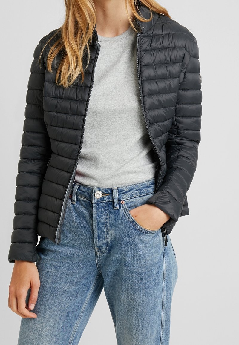 Sort quilted jakke med glat tekstur og lynlås, båret over en grå t-shirt og parret med lyseblå denim jeans.