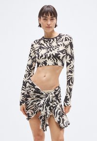 Μαύρο και κρεμ κοντό τοπ με μακριά μανίκια, διακοσμημένο με τροπικό μοτίβο φύλλων· συνοδευτική φούστα τύπου wrap με λεπτομέρεια κόμπου. Λείο, ελαστικό ύφασμα.