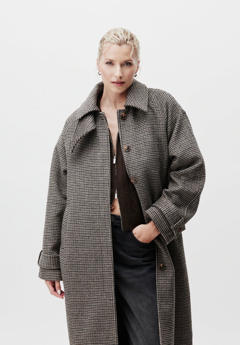 Πανωφόρι σε καφέ και μαύρο καρό, υπερμεγέθες, με μεγάλο γιακά, κουμπιά στο μπροστινό μέρος και πλαϊνές τσέπες, φορεμένο πάνω από ένα σκούρο τοπ και τζιν.