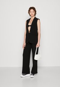 Black sleeveless blazer with a low cut, paired with black trousers. White handbag and black and white sneakers complete the outfit.