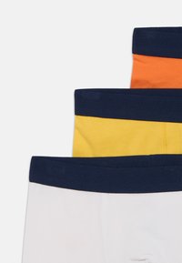 Kolm paari puuvillaseid lühikesi pükse, millel on tumesinised elastsed vöökohad; värvideks on valge, kollane ja oranž ning materjalil on sile tekstuur.