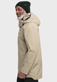 Beige wasserdichte Jacke mit Kapuze, Seitentaschen und Reißverschlussärmeln. Getragen mit einer dunklen Strickmütze, die ein dezentes Branding am Ärmel aufweist.