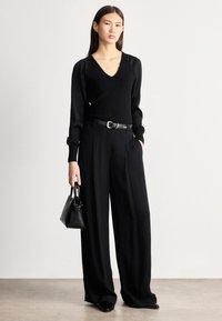 Sieviete, tērpusies melnā V veida kakla džemperī, platajiem biksēm un jostu, turot rokā mazu melnu somiņu, stāv pret vienkāršu gaišu fonu.