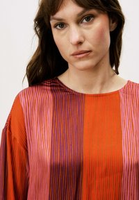 Blouse rayée rouge et pourpre avec un col rond et une coupe ample. Le tissu semble léger avec une texture lisse et un léger éclat.