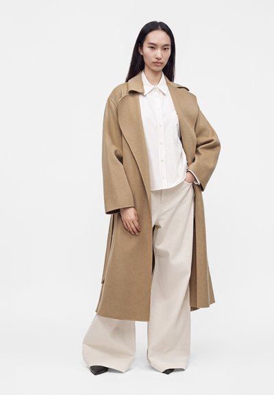 Femme portant un long manteau camel sur une chemise blanche boutonnée et un pantalon beige large, debout avec une main dans la poche devant un fond uni.