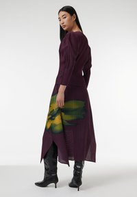Largo vestido púrpura con pliegues verticales, adornado con un gran diseño floral en verde y amarillo, combinado con brillantes botas negras hasta la rodilla.