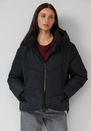 Doudoune noire avec fermeture éclair sur le devant, capuche ajustable et texture matelassée. Associée à un pull bordeaux et un jean gris.