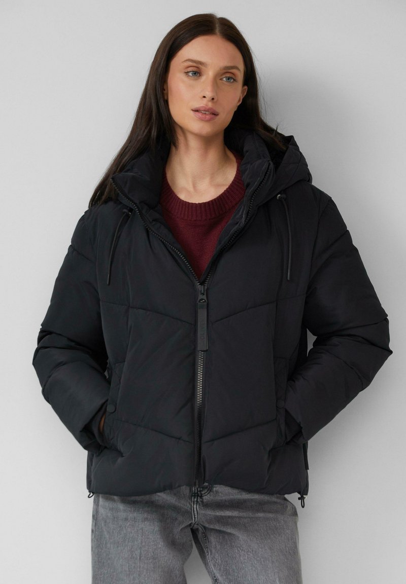 Schwarze Pufferjacke mit durchgehendem Reißverschluss, verstellbare Kapuze und gestepptem Muster. Kombiniert mit einem burgunderfarbenen Pullover und grauen Jeans.