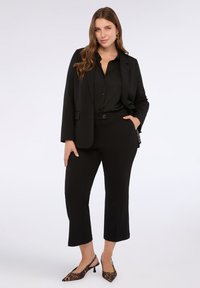 Kvinna med långt brunt hår klädd i svart kavaj, svarta croptop-byxor och slingback-skor med leopardmönster, poserar med ena handen i fickan.