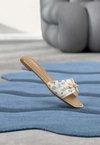 Sandale à talon plat de couleur blanc cassé avec imprimé floral. Dotée d'un accent doré et d'une semelle lisse. Conçue avec une large bride ouverte à l'avant pour le confort.