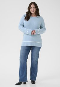 Pull en maille bleu clair à manches longues avec bordure blanche. Associé à un jean évasé bleu et des bottes noires à talons. Texture lisse.