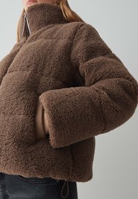 Braune Fleecejacke mit hohem Kragen und weit geschnittenen Ärmeln. Hat eine strukturierte Außenfläche, seitliche Taschen und einen körpernahen Schnitt.