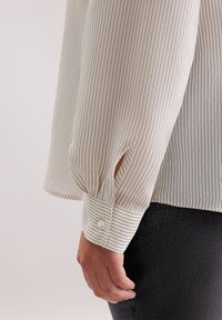 Blusa a maniche lunghe a righe color crema con strisce verticali blu. Presenta un polsino con bottoni e una fessura laterale. Tessuto testurizzato con una morbida caduta.