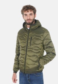 Giacca a vento imbottita verde oliva con motivo a onde trapuntate, chiusura frontale con cerniera e cappuccio. Presenta una tasca sul petto e tasche laterali con cerniere.