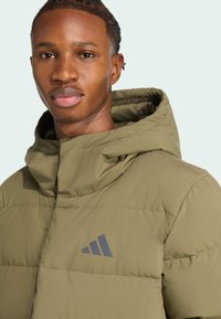 Maslinasto zelena puhovka s visokim ovratnikom i kapuljačom, glatke teksture, uzorka dijamantnog prošivanja i tamnim logotipom na prsima.