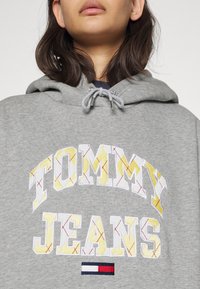 Felpa grigia con cappuccio e logo ricamato che dice "TOMMY JEANS" in bianco e giallo. Presenta un cappuccio con lacci e un accento rosso, bianco e blu.