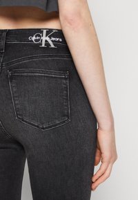 Vaqueros de mezclilla negra con bolsillos traseros y un sutil logo de Calvin Klein en la cinturilla. La tela tiene una textura clásica de mezclilla.