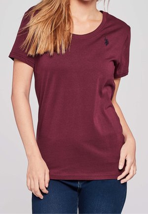 Femme portant un t-shirt bordeaux uni à manches courtes avec un petit logo noir brodé sur la poitrine gauche, associé à un jean bleu foncé.