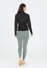 Schwarzes Langarmoberteil mit hohem Kragen, strukturierter Stoff am Rücken, kombiniert mit hellgrünen Leggings und beigen Schuhen.