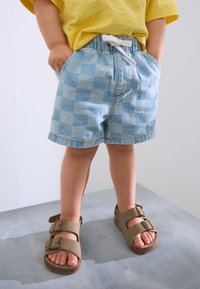 Pantaloncini a quadretti blu con fascia elastica in vita e coulisse bianca, abbinati a sandali marroni con due cinturini. È visibile una maglietta gialla a maniche corte.