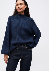 Granatowy sweter z prążkowanego dzianiny z wysokim kołnierzem, oversize'owymi rękawami i luźnym krojem, w połączeniu z ciemnoniebieskimi dżinsami. Gładka faktura, bez wzorów.