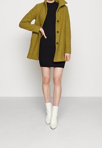Femme portant une robe noire côtelée longueur genou, un manteau boutonné vert olive et des bottines blanches, debout devant un fond uni.