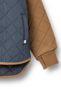 Quiltet jakke med blå og brune sektioner, der har et diagonalt diamantmønster og ribbet manchet. Inkluderer en sidelomme med diskret branding.