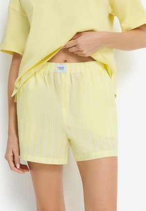 Frau trägt gelb-weiß gestreifte Shorts mit elastischem Bund und ein hellgelbes, lockeres T-Shirt, die Hand hebt das T-Shirt leicht an.