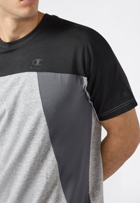 Hombre vestido con una camiseta de manga corta de Champion con bloques de color en negro, gris oscuro y gris claro, mostrando el pecho y la parte superior del brazo.