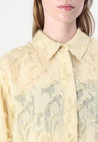 Camicia gialla trasparente con un motivo testurizzato e sfrangiato. Presenta una chiusura con bottoni sul davanti e un colletto classico. Tessuto leggero con una caduta morbida.