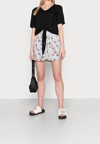 Femme portant une chemise noire à nouer, un short floral bleu clair, des sandales blanches à boucle et tenant un petit sac à main noir.