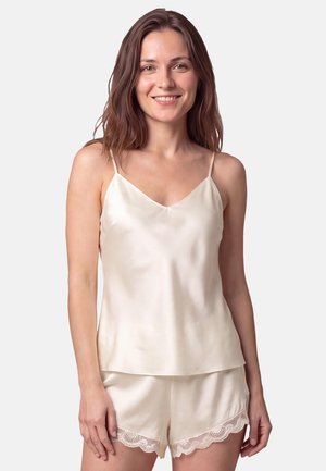 Kvinde smilende iført cremefarvet satin camisole og matchende shorts med blondekant på en ensfarvet lys baggrund.