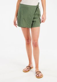 Olijfgroene wikkelrok van lichte stof met zijstrikdetail, gecombineerd met bruine sandaaltjes op blote voeten.