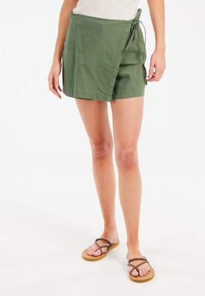 Jupe portefeuille vert olive en tissu léger avec détail de nœud sur le côté, associée à des sandales marron à lanières sur pieds nus.