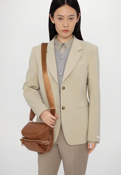 Brown leather crossbody bag with a zip pocket, chain accent, and adjustable strap, paired with a beige blazer and gray layered top.
