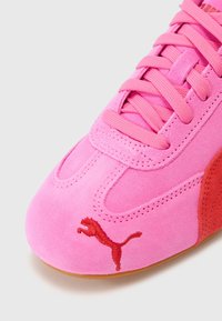 Detailní záběr na růžovou semišovou tenisku s červeným logem Puma na špičce a odpovídajícími růžovými tkaničkami na bílém pozadí.