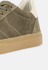 Ανδρικό αθλητικό παπούτσι από σουέτ με επάνω μέρος σε ελιάς πράσινο, μπεζ λεπτομέρειες και χοντρή σόλα από καουτσούκ σε φυσικό χρώμα. Διαθέτει ραφές στον ίδιο τόνο και στρογγυλά κορδόνια.