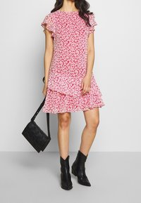 Röd blommig klänning med volangärmar och en flerlagersdesign, kombinerad med en svart axelväska och svarta ankelstövlar. Slät tyg.