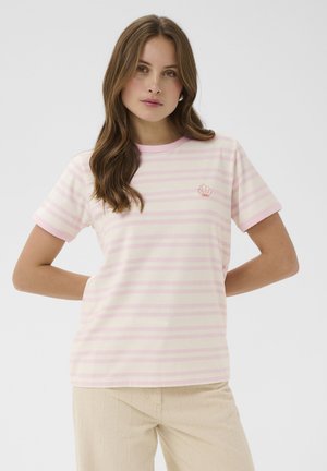 Młoda kobieta z długimi brązowymi włosami, ubrana w jasno różowy i biały paskowany t-shirt z krótkim rękawem oraz beżowe spodnie, ręce za plecami.