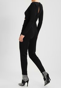 Monos de manga larga negros con un diseño retorcido y espalda descubierta. Combinado con botas metálicas de tobillo con tacón block y correa negra.