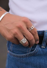 Anelli in argento con ritagli geometrici e un design a freccia incisa su una mano, che indossa un bracciale in argento texturizzato, su un tessuto di denim.