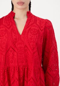 Röd bomulls klänning med broderi i ögonlapp, med djup V-ringning, långa ärmar och en rynkad midja. Strukturerade blommönster pryder tyget.
