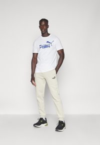 Hvid T-shirt med blå Puma-logo, beige sweatpants med logo på siden, sorte og grønne træningssko. Står foran en neutral baggrund.
