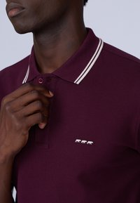 Polo bordeaux avec des rayures blanches sur le col, présentant une petite broderie blanche de trois vaches sur le côté gauche près de la patte de boutonnage.